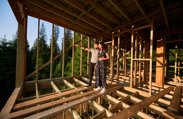 Man and woman examining their future wooden frame dwelling nestled near the forest. Youthful couple at construction site in early morning. Concept of contemporary ecological construction.