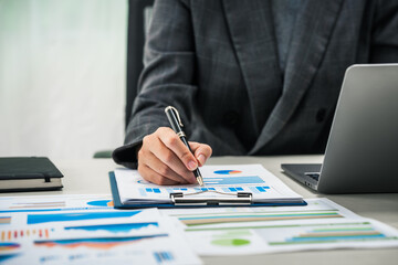 A businesswoman works diligently at her desk, reviewing financial documents and analyzing...