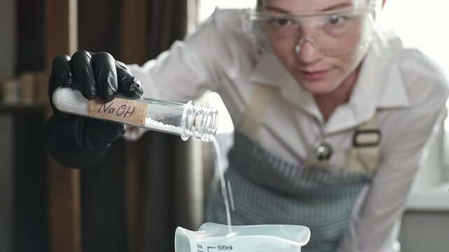A woman soap maker weighs lye on a scale to make natural soap. The master in glasses and gloves in the workshop