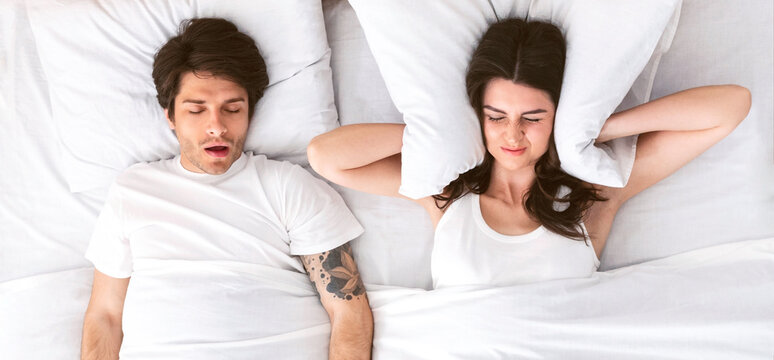 A woman lies in bed with her eyes closed and her hands covering her ears while a man sleeps beside her. The mans mouth is open as he snores loudly. The woman appears annoyed or frustrated.