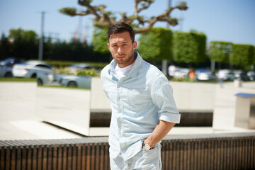 Young man in casual attire standing outdoors with a serious expression, surrounded by modern urban environment and parked cars