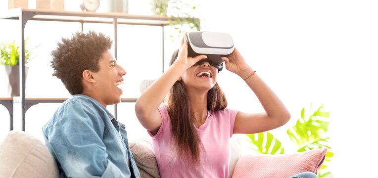 African American teen couple is enjoying a virtual reality experience indoors, with the girl wearing a VR headset and laughing as the guy watches.