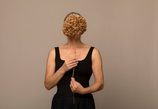 Senior woman hiding face with withered flower standing against brown background