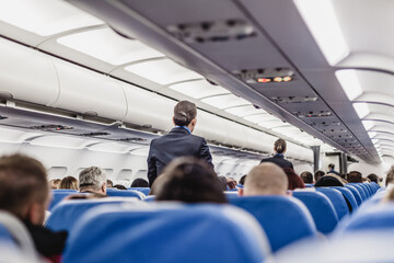 Interior of airplane with passengers on seats and stewardess walking the aisle.