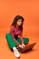 Young woman with curly hair sits on orange background in striped sweater and green pants, using laptop and staring at screen.