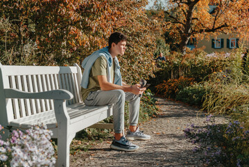 A Teenager Sits On A Bench In The Autumn Park Drinks Coffee From A Thermo Mug And Looks Into A Phone. Portrait Of Handsome Cheerful Guy Sitting On Bench Fresh Air Using Device Browsing Media Smm