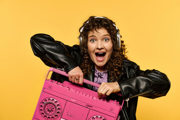 A young woman with curly hair is wearing a black leather jacket and headphones. She is holding a pink boombox and smiling.