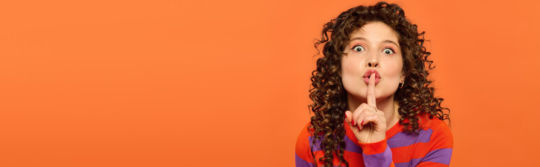 A young woman with curly hair poses against a vibrant orange backdrop, her finger pressed to her lips.