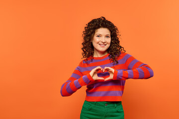 A young woman with curly hair poses against a bright orange background, forming a heart shape with her hands.