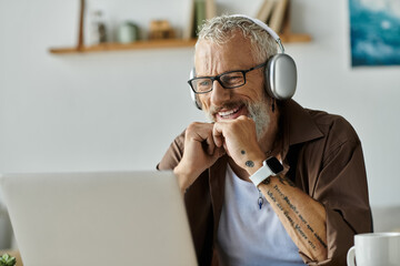 A mature gay man with tattoos and grey hair smiles while working from home on his laptop.