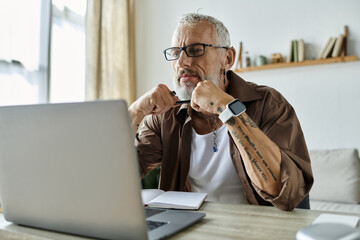 A mature gay man with tattoos and grey hair works remotely from home on a laptop.