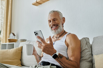 A mature man with tattoos and a grey beard smiles as he relaxes on his couch at home, scrolling through his phone.