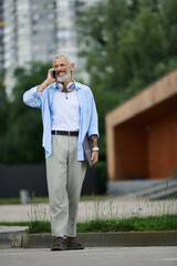 A tattooed, mature gay man with a grey beard smiles as he talks on his cell phone while walking outdoors in the city.