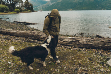 A man is petting a dog on a beach. The man is wearing a green jacket and a backpack. The dog is black and white. The beach is near a body of water