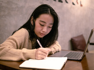 Asian woman working on a laptop in a cozy café, writing notes while focused on her task. The café's warm ambiance and rustic décor create a productive yet relaxed work environment.