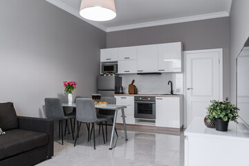 stylish interior of apartment with modern kitchen in white. kitchen design with appliances, microwave, refrigerator, stove, table, grey chairs. pink roses, fruits and wine on glass kitchen table. 