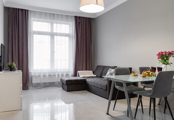 modern design of living room in apartment. Near window is big gray sofa with pillows, coffee table and plants. dining table and chairs near sofa, flowers in a vase, glasses and fruits on table
