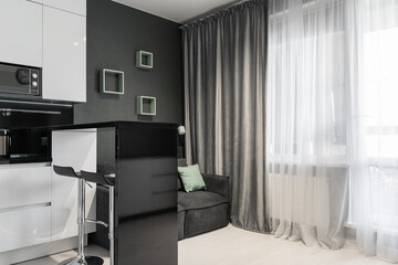 stylish interior of apartment with modern kitchen in black and white. Modern design of kitchen-living room with appliances, microwave, refrigerator, stove. grey sofa and blue cushions in living room