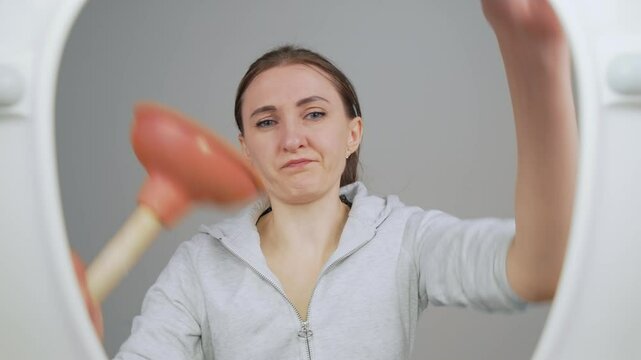Woman janitor in rubber hygiene gloves lifts lid of toilet, Female holding plunger in her hand and contemplating how to clear clog in toilet pipes. View from toilet bottom. Cleanliness and hygiene