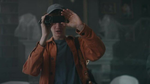 Medium shot of Caucasian guy recording content on VHS camera, filming interior of abandoned mansion while examining dark room with diverse friends