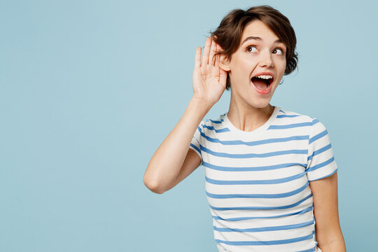 Young curious nosy woman wears striped t-shirt casual clothes try to hear you overhear listening intently isolated on plain pastel light blue cyan color background studio portrait. Lifestyle concept.