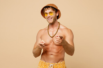 Young sexy man wear yellow shorts swimsuit hat relax rest near hotel pool point finger camera on you motivating encourage isolated on plain beige background. Summer vacation sea rest sun tan concept.