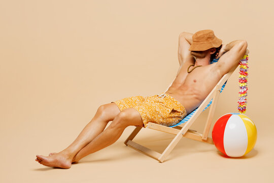 Full body young sexy man wear yellow shorts swimsuit relax rest near hotel pool sit in deckchair cover head face with hat isolated on plain beige background. Summer hot vacation sea sun tan concept.