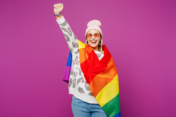 Young fun happy lesbian woman wrapped in rainbow flag wear pink animal clothes raise up hands clench fist isolated on plain purple background studio portrait. Pride day June month love LGBTQ concept.