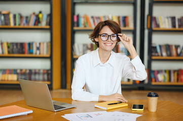 Young smiling happy cheerful fun successful employee business woman wearing white shirt casual clothes touching glasses look camera sit work at office desk with pc laptop. Achievement career concept.