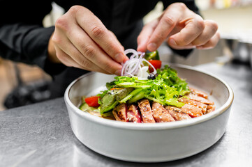 Close-up of hands garnishing a fresh salad with grilled chicken, vegetables, and greens in a white bowl.