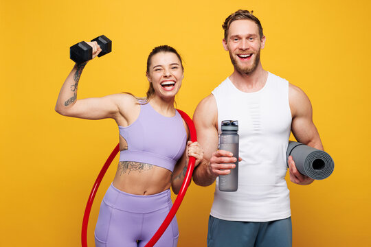 Young happy fitness trainer instructor sporty two man woman wearing blue clothes spend time in home gym hold yoga mat dumbbells hula hup isolated on plain yellow background. Workout sport fit concept.