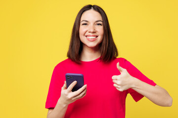 Naklejka premium Young smiling happy cheerful woman she wear pink t-shirt casual clothes hold in hand use mobile cell phone show thumb up isolated on plain yellow orange background studio portrait. Lifestyle concept.