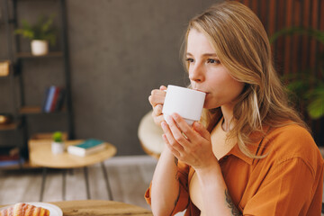Close up side view young woman wears orange shirt drink coffee sitting alone at table in coffee shop cafe relax rest in restaurant during free time indoors. Freelance mobile office business concept