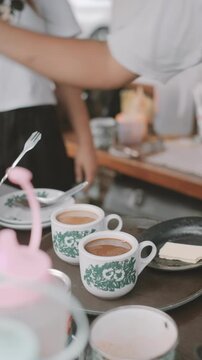 Pouring coffee from a Singaporean traditional coffee pot into vintage-style mugs and stirring it. Kopitiam coffee style. Bokeh foreground and background.