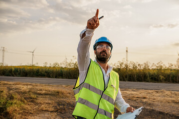 Maintenance engineer points with his hand at a defective turbine in a wind turbine park. Portrait of skilled professional. European green deal