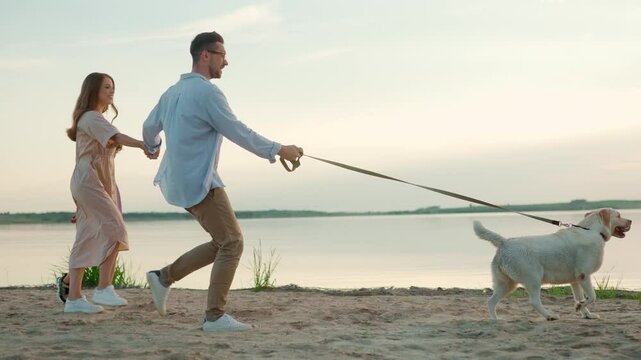 Caucasian family walking their dog together next to large lake. Father, mother and son actively spending their time. Wonderful beach catching light from sunset. Dog rushing in front of family.