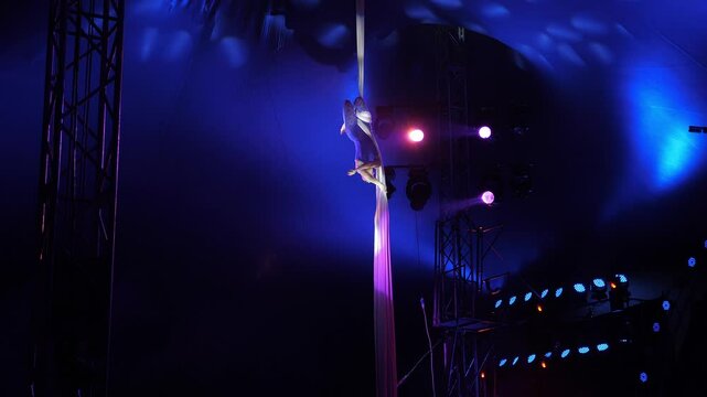 A flexible aerial gymnast performs a dangerous trick on fabric high under the dome of the circus, she rotates under the light of flashing multicolored spotlights. A circus show. An aerial acrobat.