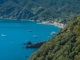 Fototapeta premium Picturesque distant view of Monterosso al Mare with beach, ships and boats seen from Azure Trail to Vernazza with green cliffs, rock, clear blue sky and sea. National park Cinque Terre, Liguria, Italy