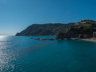 Fototapeta premium Picturesque distant view of Monterosso al Mare with beach, ships and boats seen from Azure Trail to Vernazza with green cliffs, rock, clear blue sky and sea. National park Cinque Terre, Liguria, Italy