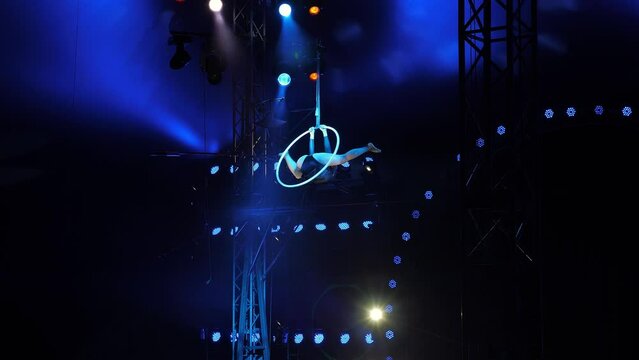 A flexible aerial acrobat performs with a hoop under the dome of the circus, she shows various tricks under the light of blue spotlights. Circus show with aerialists and acrobats.