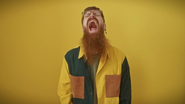 Frustrated young redhead man in glasses screams in fury, enraged and furious, showcasing aggression and rage over a yellow isolated background