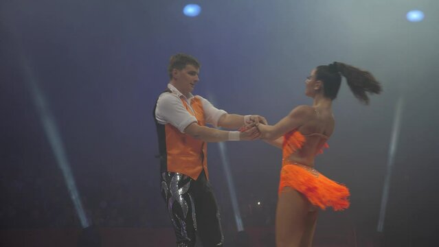 A pair of acrobats on roller skates in beautiful stage costumes perform in the circus arena, spinning around themselves on a small stage. Concert lighting.