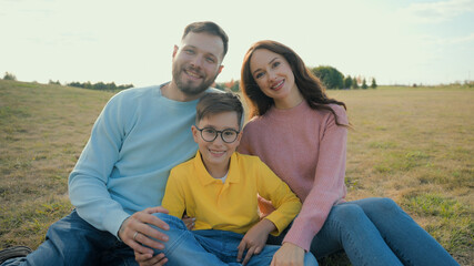 Cute Caucasian happy family portrait joyful man male dad smiling woman female mom little boy child kid son sitting together hug embrace looking camera outside nature field farmland park picnic nature