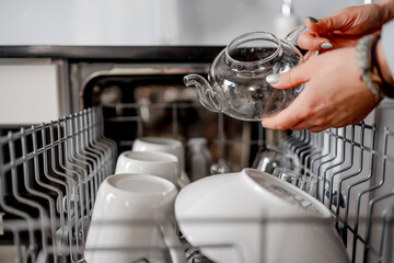 Female Hands Hold A Transparent Teapot Opposite A Dishwasher Arranging Clean Dishes