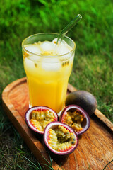 Passion fruits cut in half next to a glass of juice with ice on a wooden tray against a background of green grass