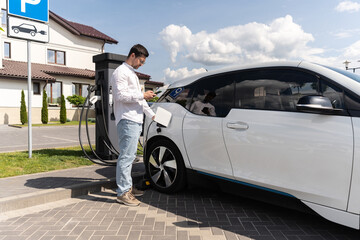 Man Charging Electric Car At Station With Smartphone On Sunny Day