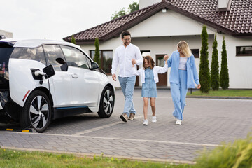 Happy Family Walking Near Electric Car And Modern House
