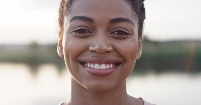 Close front view of African American woman face. Girl looking to side and afterwards directly into camera. Smiling with very white teeth. Stable water of large pond of lake in background.