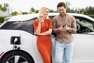 Smiling Couple Charging Electric Car While Using Smartphone. Man And Woman Standing By EV, Enjoying Coffee Break. Modern Technology, Sustainable Transportation, Eco-Friendly Lifestyle.