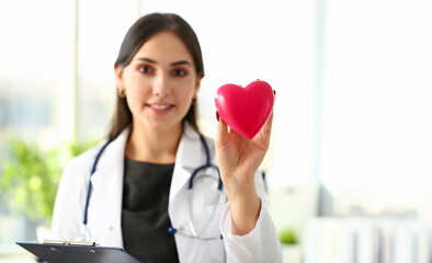 Beautiful smiling blond female doctor hold in arms red toy heart closeup. Cardio therapeutist...
